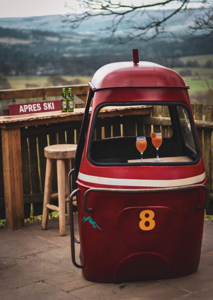 The gondola is placed on a stone patio, offering a scenic countryside view with rolling hills in the background. A wooden bar area, complete with rustic stools, sits nearby, adorned with a sign reading "APRES SKI" and a couple of green beer bottles. Two elegant glasses filled with a vibrant orange cocktail, garnished with dried fruit, rest inside the gondola, adding a touch of warmth and indulgence to the scene.
